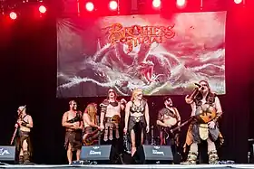 A photo of seven people on a stage, all dressed as Vikings, holding instruments like microphones and guitars, with drums in the back. There is a poster of their album Emblas Saga in the background.