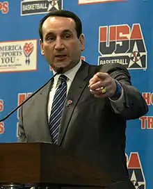 A head shot of a middle-aged Caucasian man with dark black hair is smiling. He is wearing a dark gray suit with a white undershirt and a blue tie.